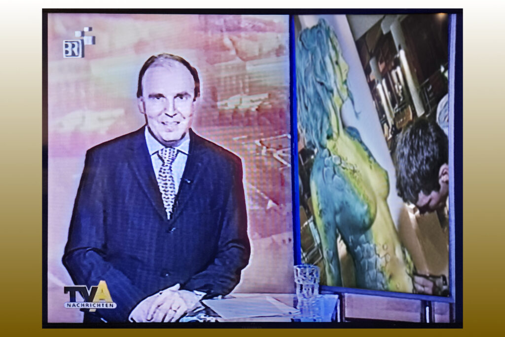Ein Fernsehmoderator steht vor einem Bildschirm, auf dem Künstler Jörg Düsterwald zu sehen ist, während er ein nacktes Fotomodell bemalt.