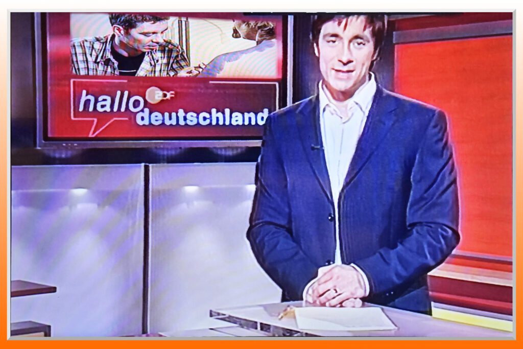 Ein Fernsehmoderator steht vor einem Bildschirm, auf dem Künstler Jörg Düsterwald zu sehen ist.