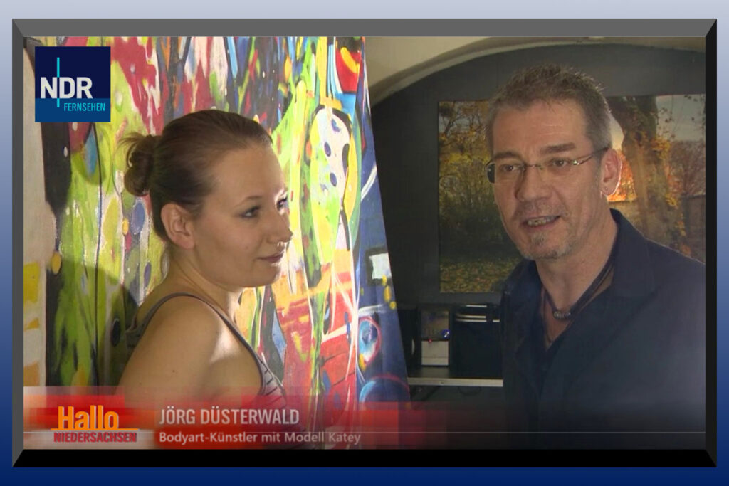 Künstler Jörg Düsterwald steht zusammen mit einem Fotomodell vor einer bunten Leinwand. Das Fernsehen berichtet über eine Kunstaktion.