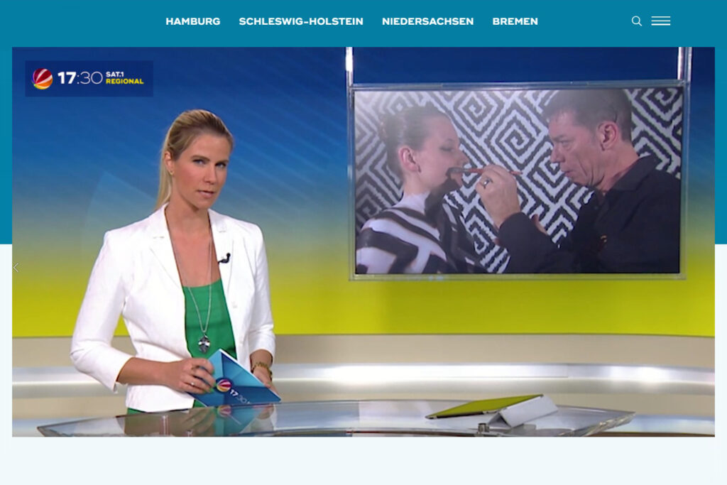 Eine Fernsehmoderatorin steht vor einem Bildschirm, auf dem zu sehen ist, wie Künstler Jörg Düsterwald einem Fotomodell das Gesicht bemalt.