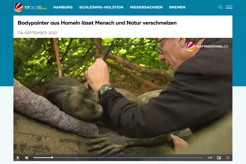 Künstler Jörg Düsterwald bemalt in einem Wald ein Fotomodell so mit Farbe, dass die Frau zur Umgebung passt.