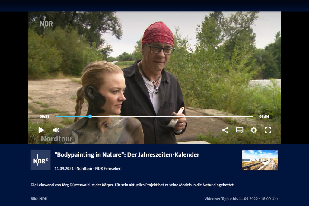 Künstler Jörg Düsterwald steht mit einem Fotomodell am Waldrand und wird von einem Fernsehteam interviewt.