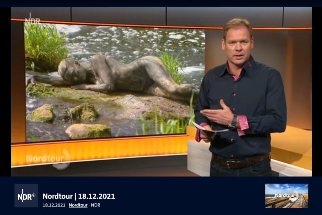 Der Moderator einer Fernsehsendung zeigt auf einen Bildschirm im Hintergrund. Darauf ist eine von Künstler Jörg Düsterwald bemalte Frau zu sehen, die in einem Fluß auf einem Stein liegt.