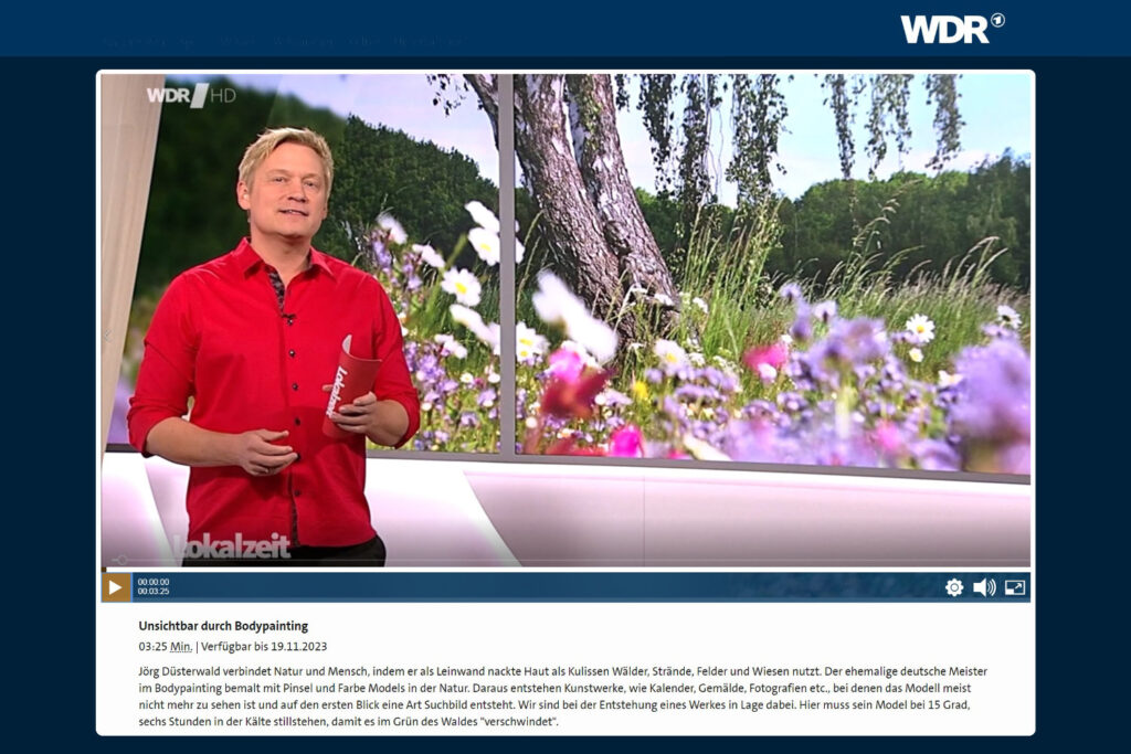 Der Moderator einer Fernsehsendung steht vor einem großen Bildschirm im Hintergrund. Darauf ist eine von Künstler Jörg Düsterwald bemalte Frau zu sehen, die in in einer Blumenwiese an einem Baum lehnt.