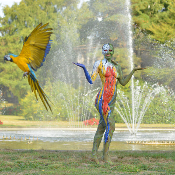 Ein Fotomodell wurde für das Kunstprojekt ANIMAL ART von Künstler Jörg Düsterwald vollständig bunt bemalt. Die Frau posiert mit Papageien. Ein Vogel fliegt davon.