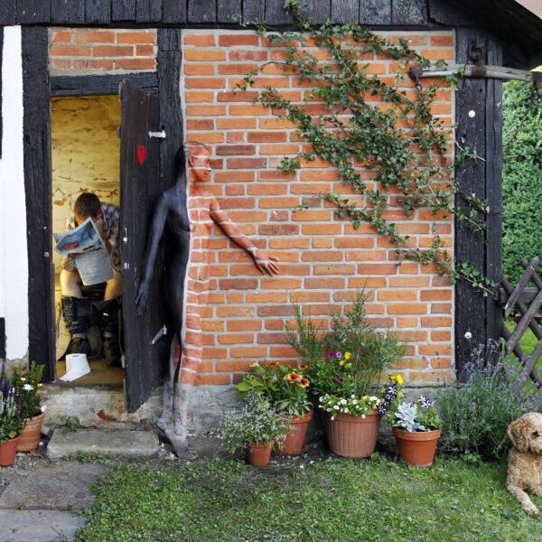 Für das Kunstprojekt DOOR ART ließ sich ein nacktes Fotomodell von Künstler Jörg Düsterwald vollständig so mit Farbe bemalen, dass es genau zur Umgebung von Türen und Toren passt. Sie steht an einer Stinmauer neben einem Plumsklo, auf dem ein Mann sitzt und Zeitung ließt. Ein Hund sitzt daneben.