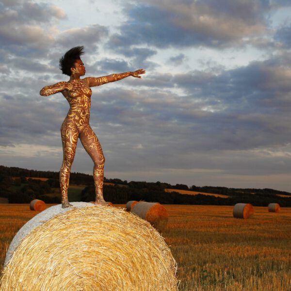 Für das Kunstprojekt AGRAR ART hat Bodypaint-Künstler Jörg Düsterwald mit Körperfarbe ein dunkelhäutiges Fotomodell mit goldfarbenen Ornamenten bemalt. Die Frau posiert in der Abendsonne bei Strohballen auf einem Strohfeld.