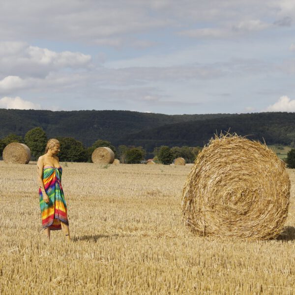 Im Rahmen des Kunstprojektes AGRAR ART hat Bodypaint-Künstler Jörg Düsterwald ein nacktes Modell vollständig so mit Körperfarbe bemalt, dass die Frau zur landwirtschaftlichen Fläche passt, in der sie sich aufhält. Sie befindet sich zur Erntezeit auf einem Strohfeld neben großen, runden Strohballen.