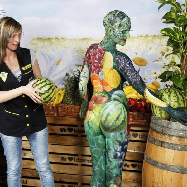 Für das Kunstprojekt WORKING ART, bei dem Berufe mit Bodypaintingmodellen dargestellt werden, hat Künstler Jörg Düsterwald ein Fotomodell vollständig bemalt. Die mit Obst und Früchten gestylte Frau hält sich zusammen mit einer Verkäuferin an einem Obststand auf.