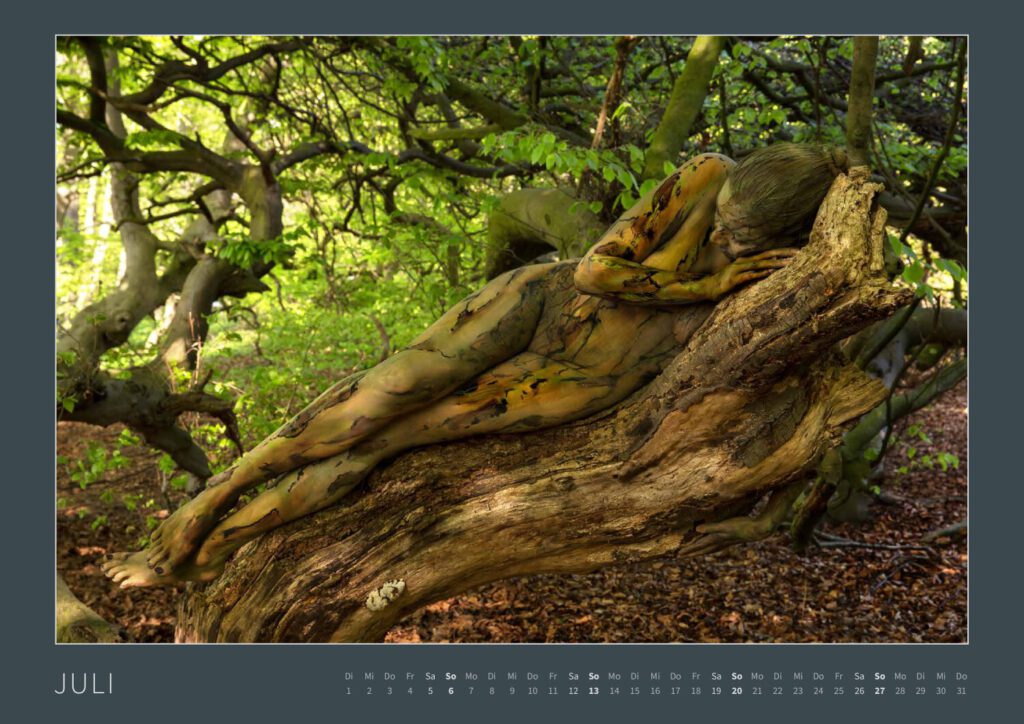 Das Monatsblatt Juli vom Bodypainting-Kalender NATURE ART 2025 von Künstler Jörg Düsterwald. Ein bemaltes Fotomodell liegt seitlich auf einem geschwungenen Baumstamm.