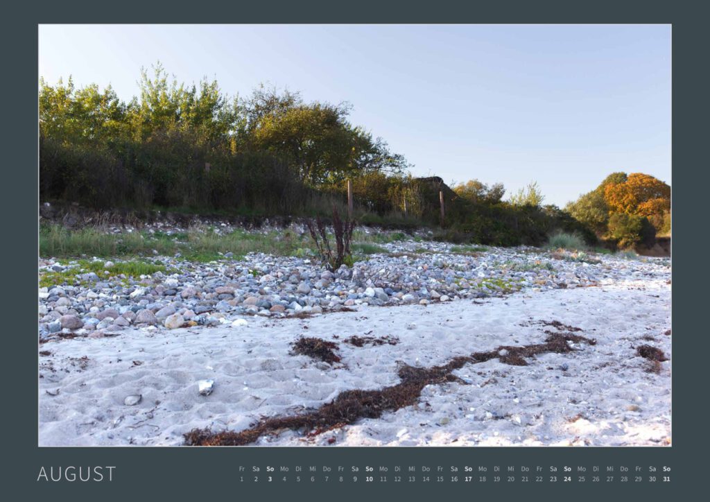 Das Monatsblatt August vom Bodypainting-Kalender NATURE ART 2025 von Künstler Jörg Düsterwald. Ein bemaltes Fotomodell liegt zwischen vielen kleinen Steinen an einem Strandabschnitt.