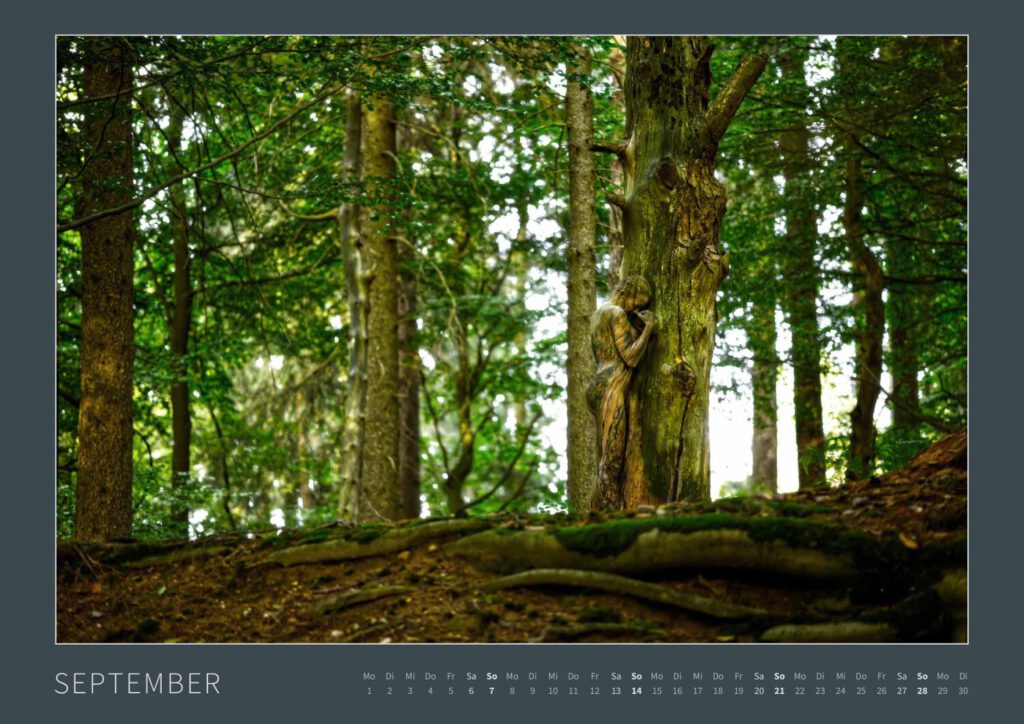 Das Monatsblatt September vom Bodypainting-Kalender NATURE ART 2025 von Künstler Jörg Düsterwald. Ein bemaltes Fotomodell steht an einem rustikalen Baumstamm im Wald mit grünen Blättern.