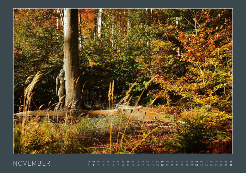 Das Monatsblatt November vom Bodypainting-Kalender NATURE ART 2025 von Künstler Jörg Düsterwald. Ein bemaltes Fotomodell lehnt an einem Baumstamm in einem herbstbunten Wald.