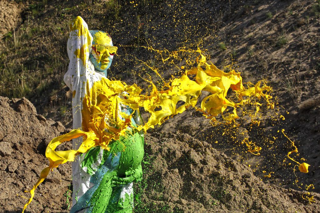 Für das Outdoor-Projekt SPLASH ART hat Künstler Jörg Düsterwald ein schwangeres Fotomodell mit Körperfarbe bemalt und anschließend in einem Sandabaugelände mit flüssiger Farbe übergossen.